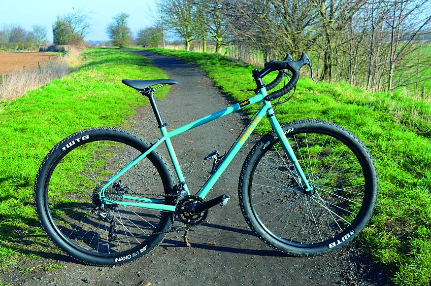 Genesis Vagabond, a turquoise adventure/gravel bike with drop bars, propped up on a gravel path