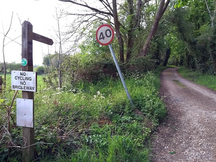 A landscape photo leading into a gravel path, sheltered by trees. A wooden signpost reads 'Public footpath', with an additional notice of 'NO CYCLING NO BRIDLEWAY'