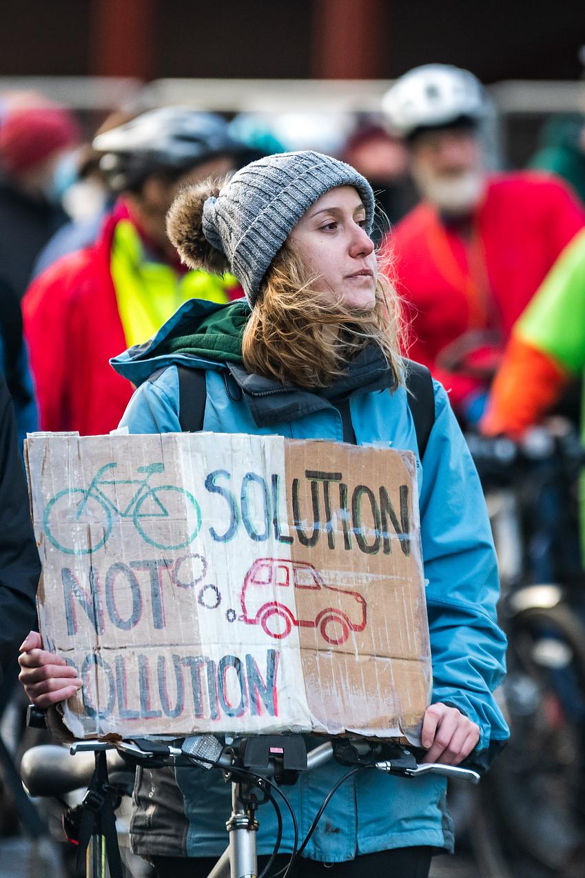 A women stands with her bicycle, holding a placard calling for cycling as a solution to car generated pollution