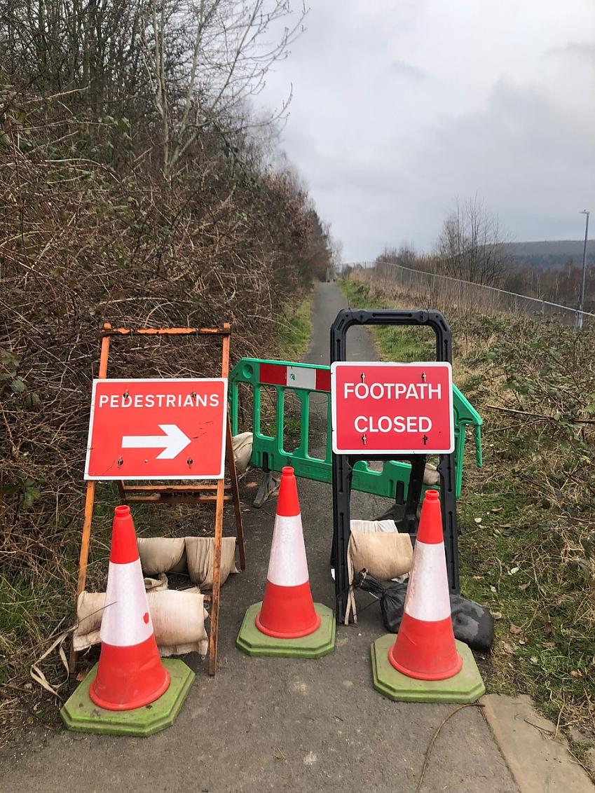 A greenway path is blocked by cones and barriers. Signage requests path users to take an alternative route