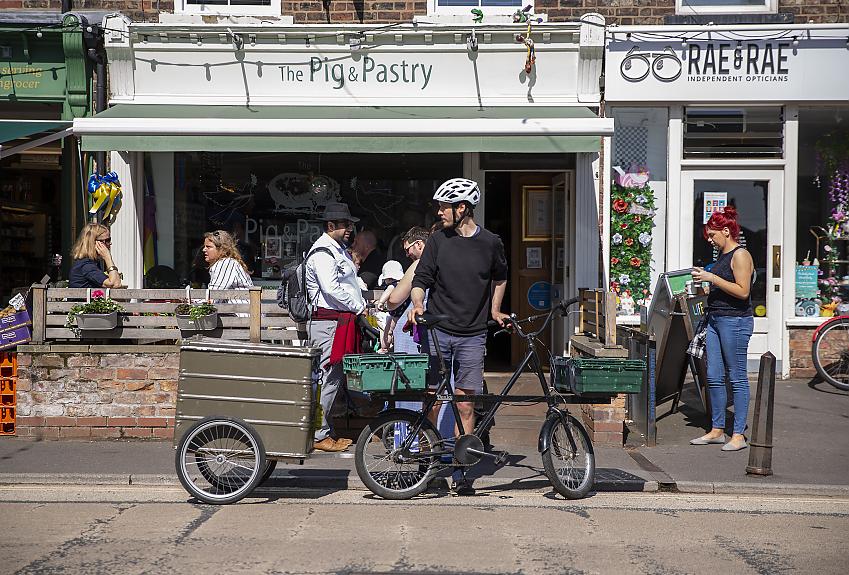 A male cyclist stands outside an al fresco dining area of a cafe with this electric cargo bike, a trailer is attached
