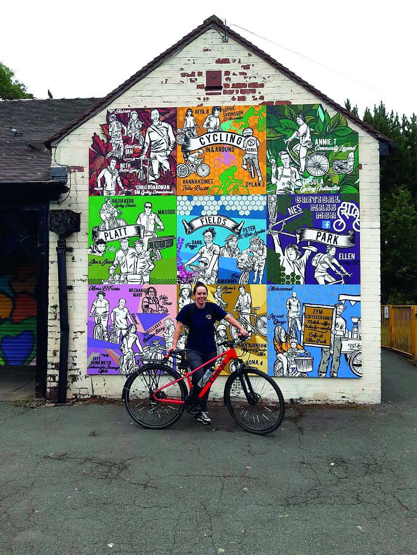A female stands with her bicycle in-front of a painted mural. The colourful mural shows important figures in Manchester's cycle scene
