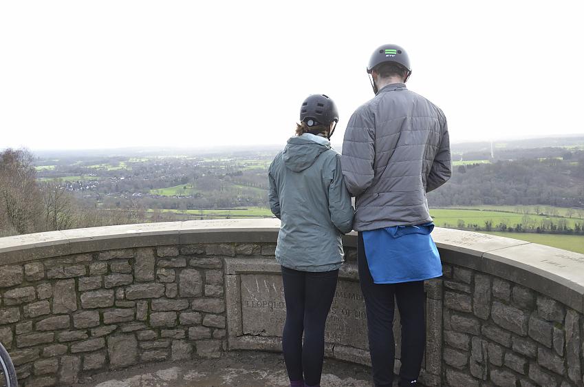 Two people in cycling kit are at the top of Box Hill and looking out over the viewpoint to the landscape laid out before them
