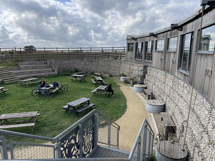A long shot of the outside space at Yorkshire Wildlife Trust Discovery Centre, with picnic tables on a grass oval. Two tables are occupied. One person is sitting on their own, while another sits with two bikes