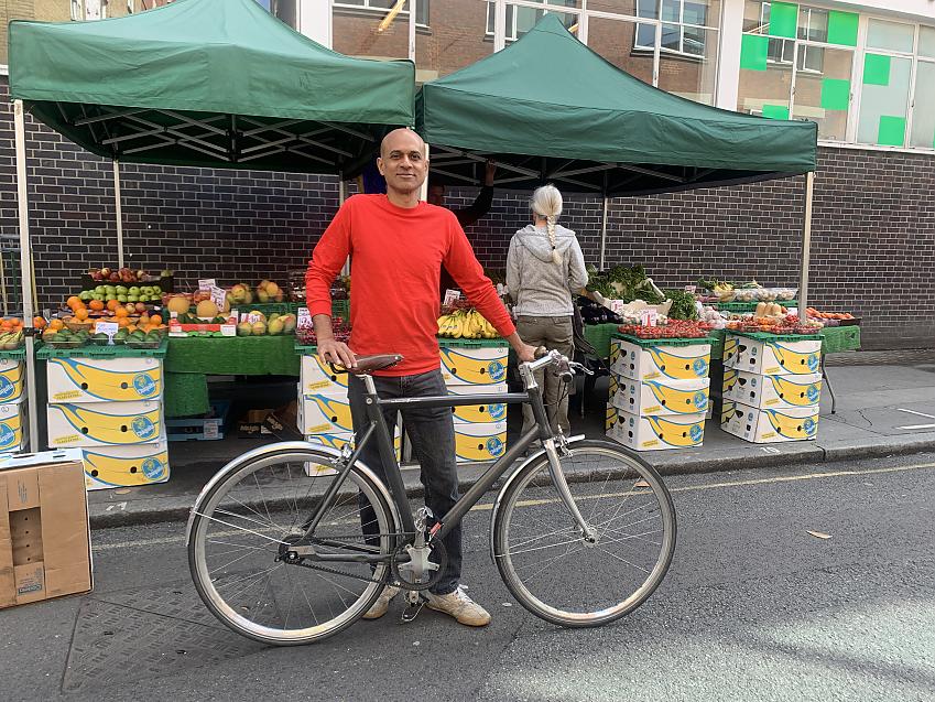 A man is standing with his grey fixie bike in front of a fruit and vegetable stall in a London street
