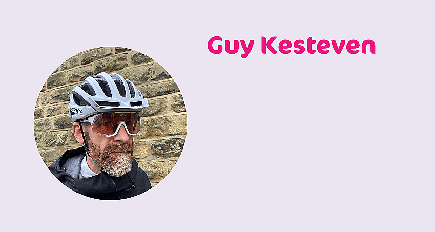 A headshot of Guy Kesteven. He is wearing a grey cycle helmet, cycling glasses and black waterproof. He has a beard. His name is written in pink alongside the image, which is in a circle.