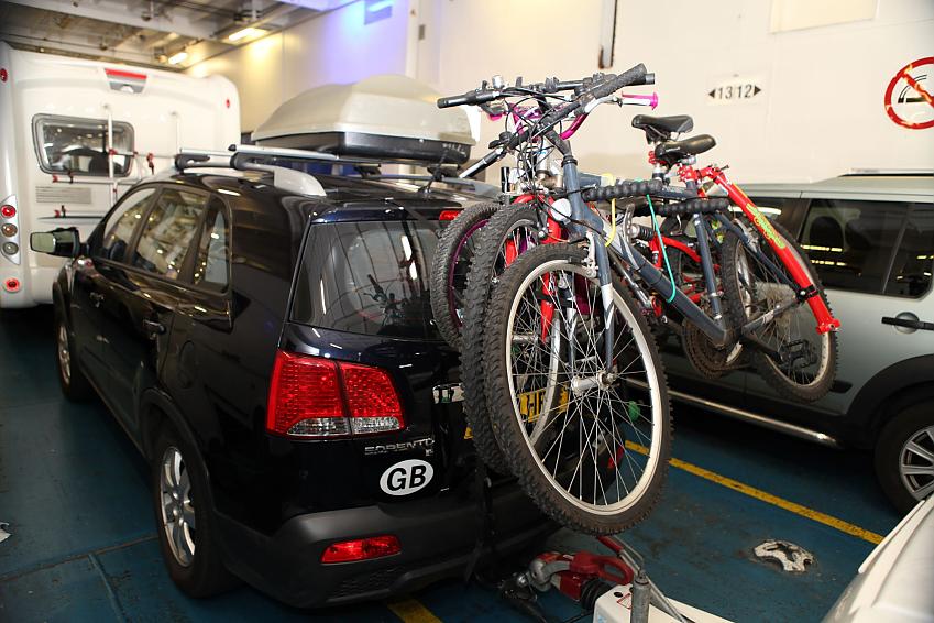 A dark blue car is parked in the car hold of a ferry. There are three mountain bikes on a rear-mounted rack on the car
