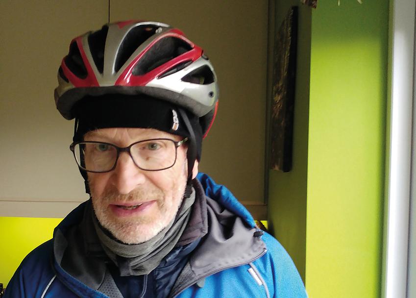 A headshot of Andrew Venables. He is wearing a silver and red cycle helmet and blue waterproof. He has glasses and a grey beard