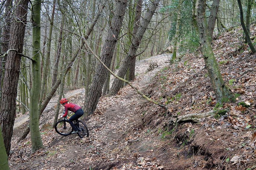 A man is riding a black mountain bike down a forested slope. He is wearing mountain bike kit and a helmet