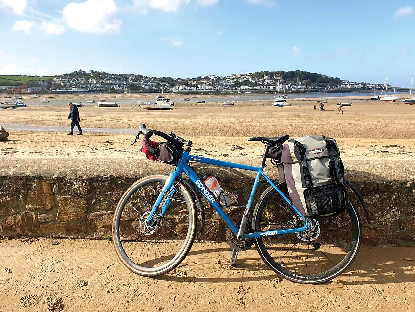 A blue Sonder touring bike is leaning against a seawall. In the background is the beach with a man walking on it and boats on the sand