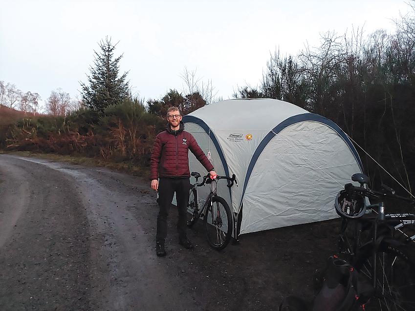 A man is standing next to a domed tent. He is wearing glasses, a warm padded jacket and trousers. He is holding a silver mountain bike