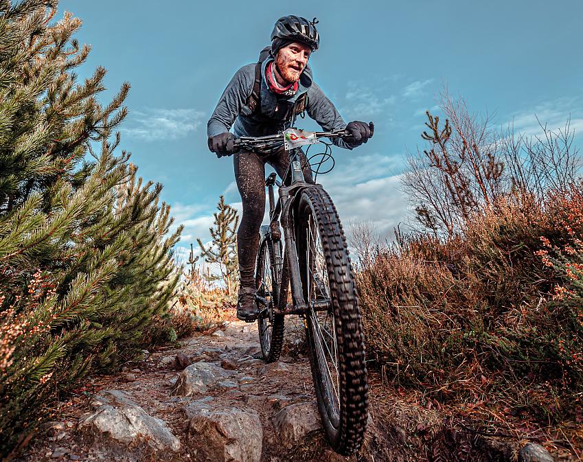 A man riding a mountain bike through rough terrain. He's wearing cycling leggings and a warm jersey; he is very muddy