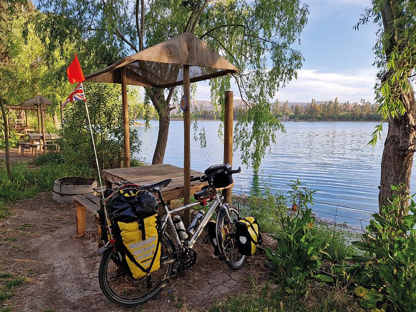 A loaded up touring bike is leaning against a wooden table with a canopy over it. There is a wide river in the background and lots of greenery