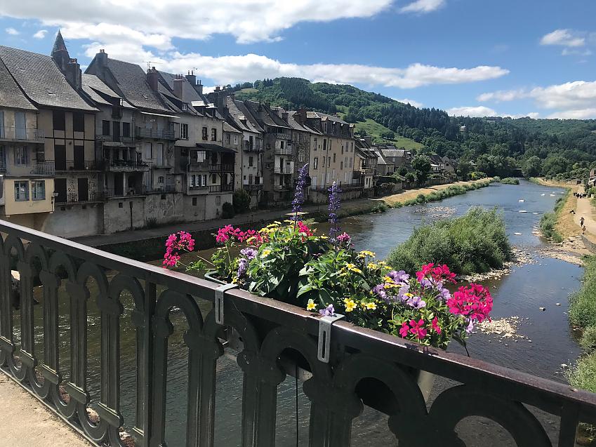 Taken over the railings of a bridge, you can see a river wending its way into mountains. There is a row of old-looking houses on the bank and planter of flowers has been attached to the railings
