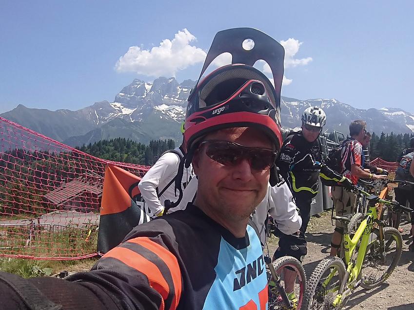 A man is taking a selfie just before setting off on a ride. He's wearing cycling kit. There are more riders in the background and behind them are the Alps