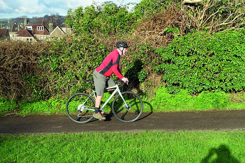 A man is riding a matt green road bike along a paved lane. He is wearing cargo shorts and a red and black long-sleeved top and a cycle helmet