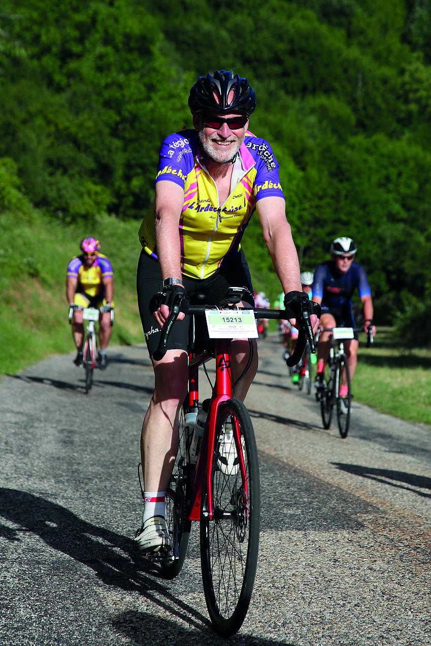 A man is riding towards the camera on a red road bike. He is wearing cycling kit, helmet and sunglasses. He's smiling. An event number on the front of his bike reads 15213.