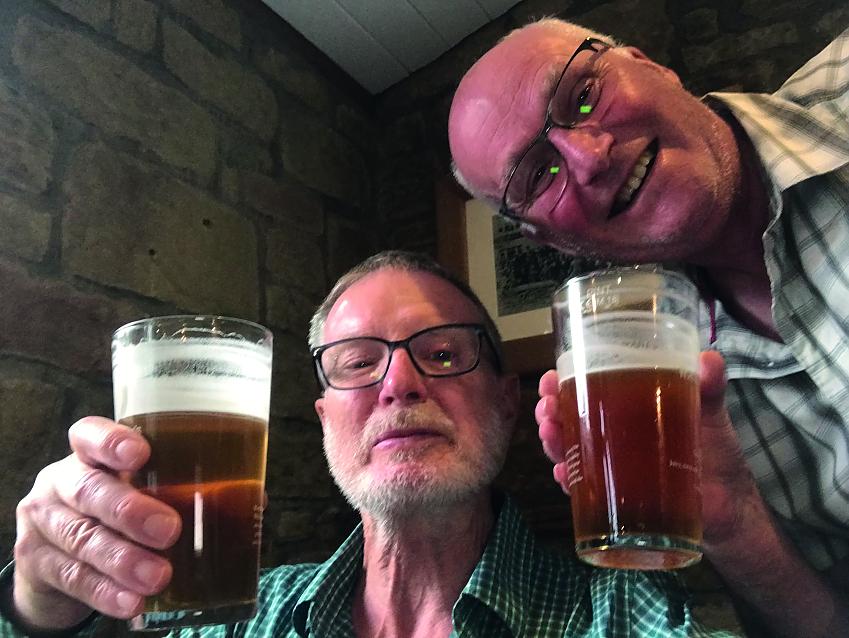 Two men are holding up pints of beer to the camera. They're smiling and are both wearing green check shirts and glasses.