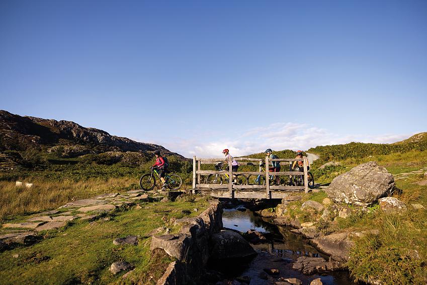 Four cyclists are cycling across a wooden bridge over a stream through mountains