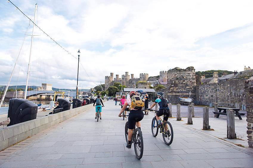 A group of cyclists on mountain bikes are riding along a shared path towards a castle