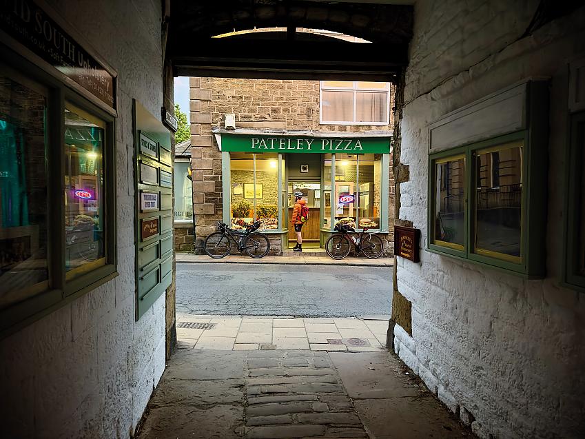 Looking through an alleyway to Pateley Pizza shop, with a cyclist standing in the doorway and a bike to either side of the door