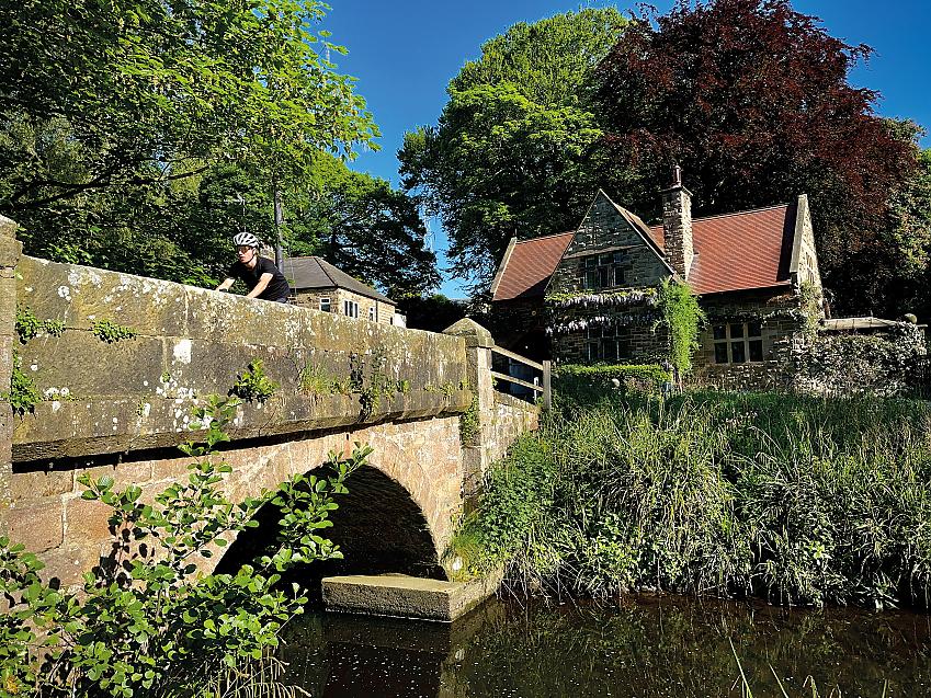 A man in a black T-shirt, shades and helmet is cycling across a stone bridge