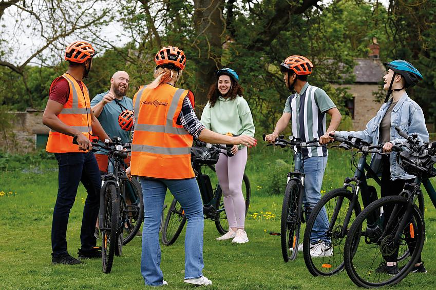 A group of people is standing in a field. They are holding bikes and wearing normal clothes and helmets. A woman is standing at the front without a bike and looks like she is explaining something