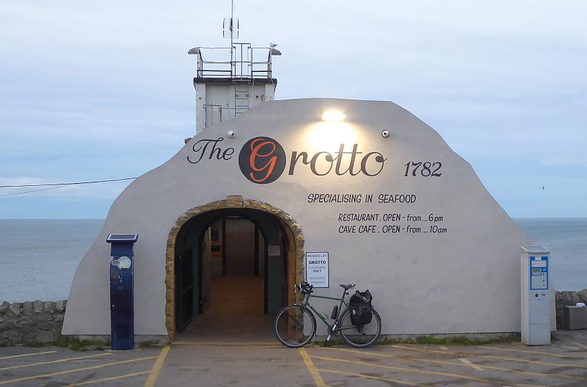 A white building is outlined against the sea in the background. It bears the writing The Grotto 1782, specialising in seafood. It also has the opening times. A packed touring bike is leaning against it. The door is open and it leads to a lift