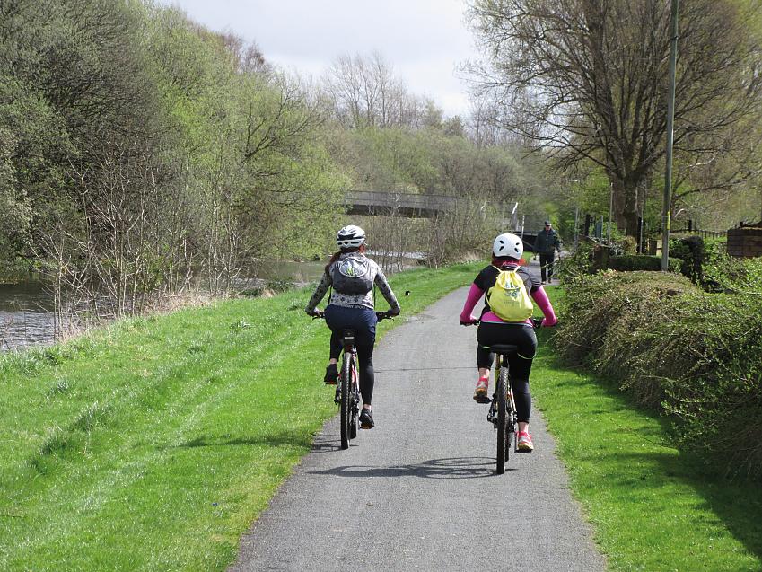 Two cyclists are riding along a paved path through countryside. There is a canal running alongside. They are wearing backpacks and cycle helmets
