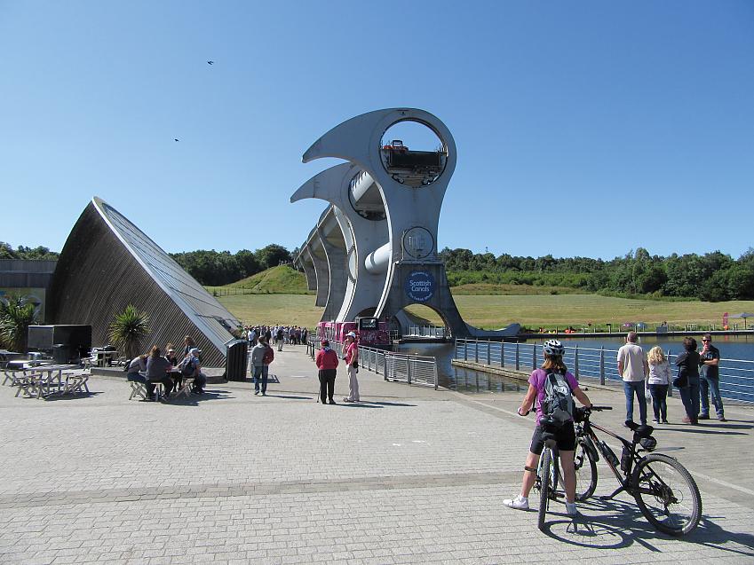 A large boat lift in the canal. There is a cyclist in the foreground standing astride a bike and holding another one, she's looking up at the boat lift. There are also lots of people sitting at tables or walking past