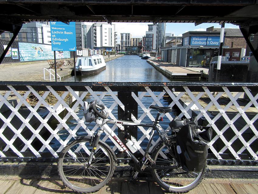 A laden touring bike is leaning against a bridge over a canal. There's a narrowboat on the canal and you can see a sign that reads Welcome to Lochrin Basin at Edinburgh Quay