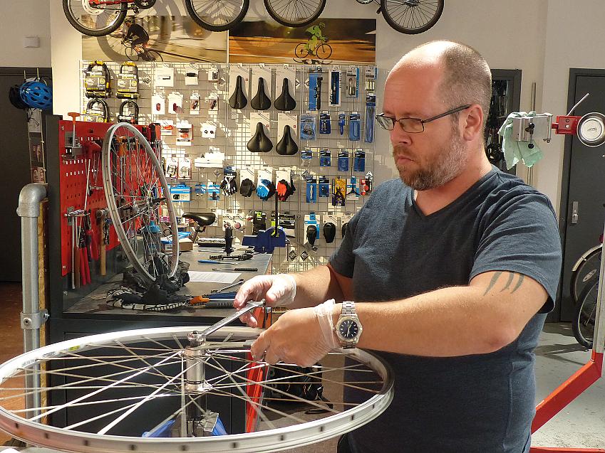 A man with a beard is adjusting a bicycle wheel. He's in a cycle workshop and in the background are lots of cycling accessories