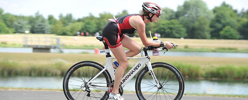 A woman is cycling on a racing cycle with triathlon bars. She's wearing a skin suit. She's cycling alongside a reservoir.