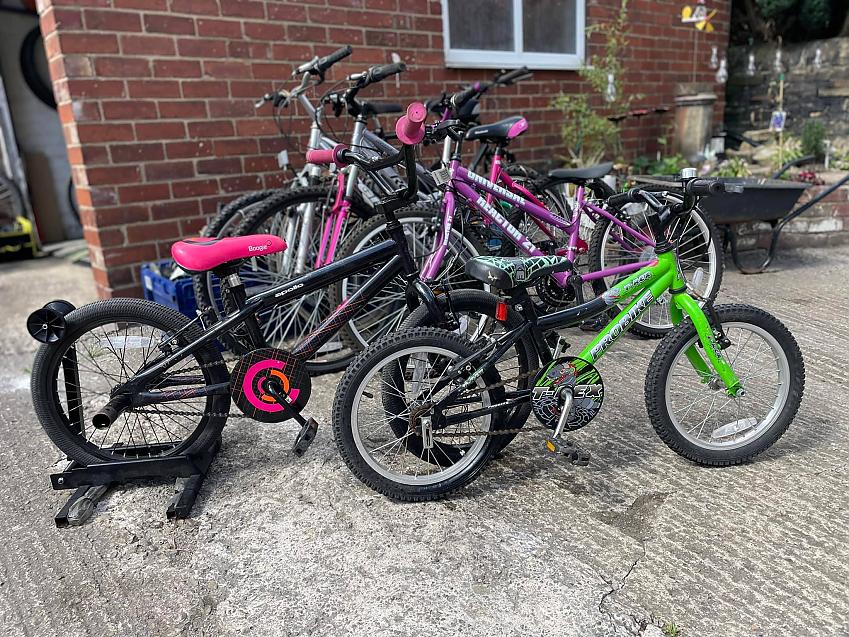  a pink and black one for a six-year-old and green and black one for a four-year-old are on a stand in front of lots more bikes leaning against the wall of a garage