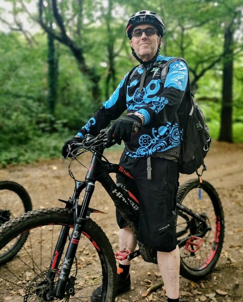 A man is standing astride a mountain bike in a forest. He's wearing black shorts and a COG Cycling club jersey, helmet and sunglasses. He's smiling