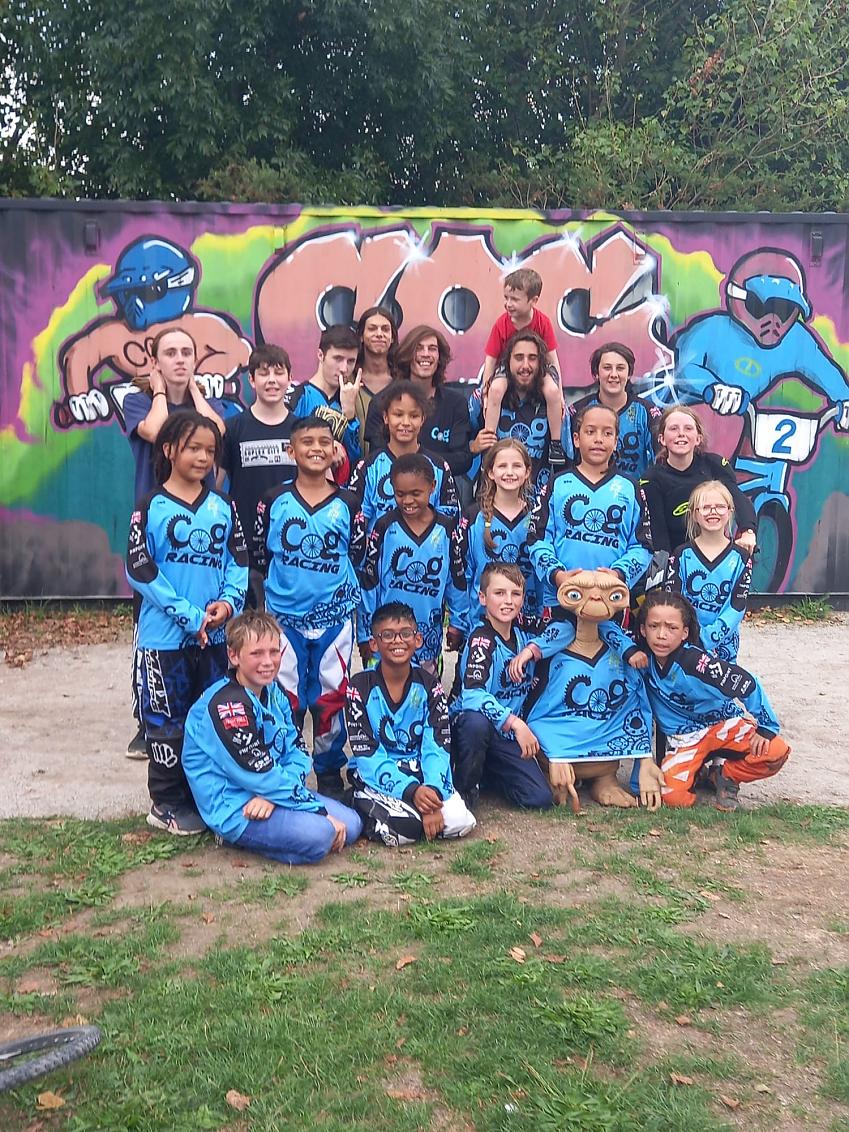 A group of children is posing with an ET figure. They are all wearing COG Cycling club jerseys. In the background is a shipping container which has been decorated with a graffiti-style image showing the club name and two people racing on BMXes