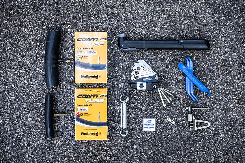 Some cycling maintenance tools are laid out on a pavement, including a bicycle pump, multitool, spare inner tubes, tyre iron and spanners