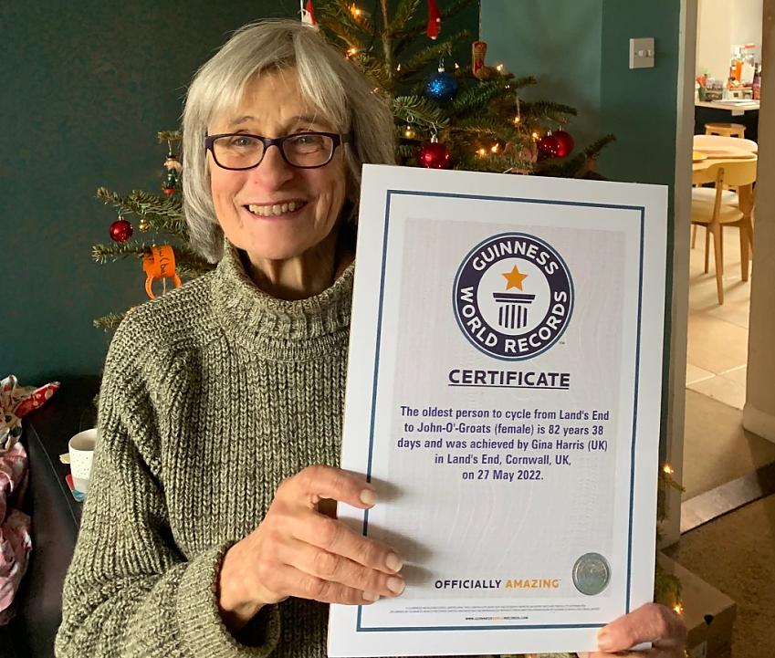 A woman with grey hair and glasses is holding up a Guinness World Records certificate showing that she is officially the oldest woman to cycle Land's End to John o'Groats. It's Christmas and there is a decorated tree behind her