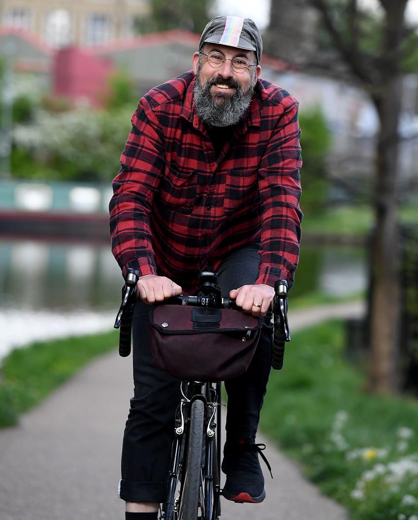 A man is riding a bike towards the camera. He's smiling. He's wearing a cap, checked shirt, black trousers and trainers. He has a beard and glasses