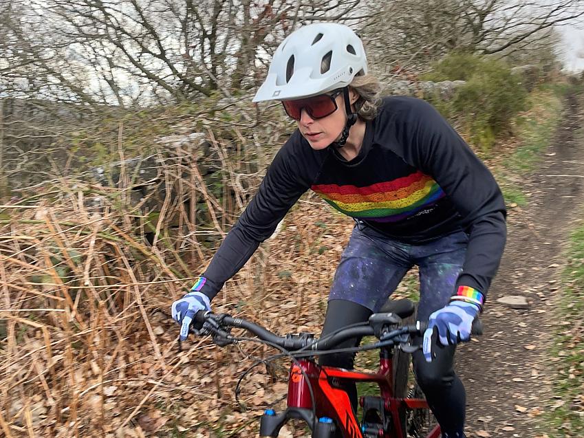 A woman is riding a mountain bike downhill. She's wearing a helmet and sunglasses, black jersey with rainbow stripes, night sky shorts and black leggings. She looks like she's concentrating hard