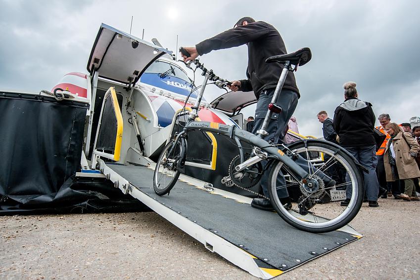 A man is wheeling a grey folding bike onto a Hovertravel vessel. He is wearing grey cargo pants and a black fleece. There are some people in the background queuing to get onto the hovercraft