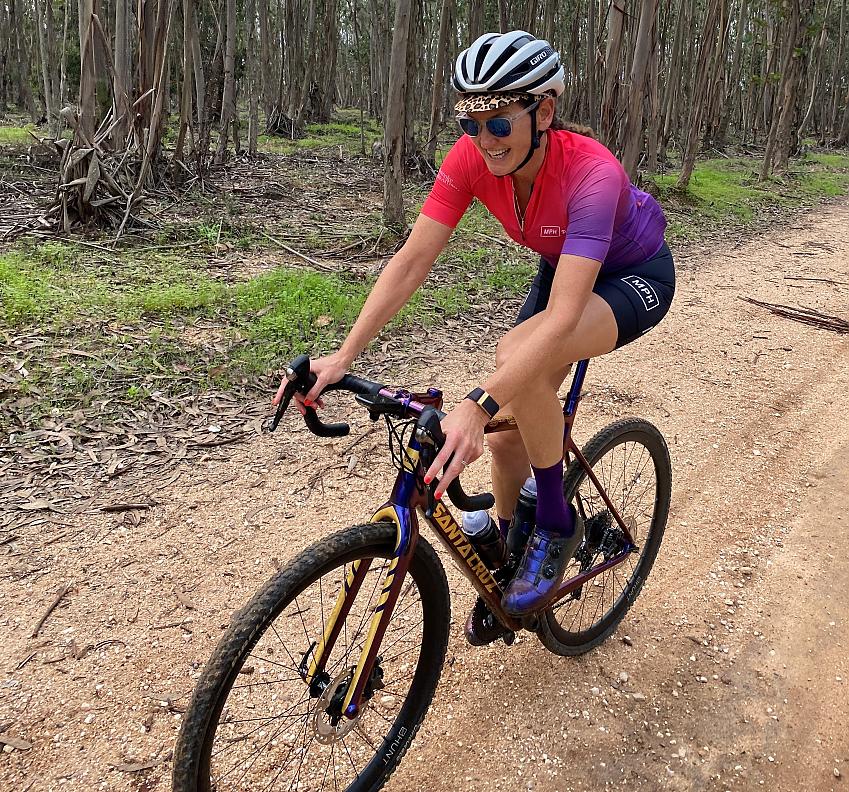 A woman is cycling on a mountain trail. She is wearing bright cycling gear and a helmet, an animal print cap can just be seen under her helmet. She's smiling. She's on a gravel bike