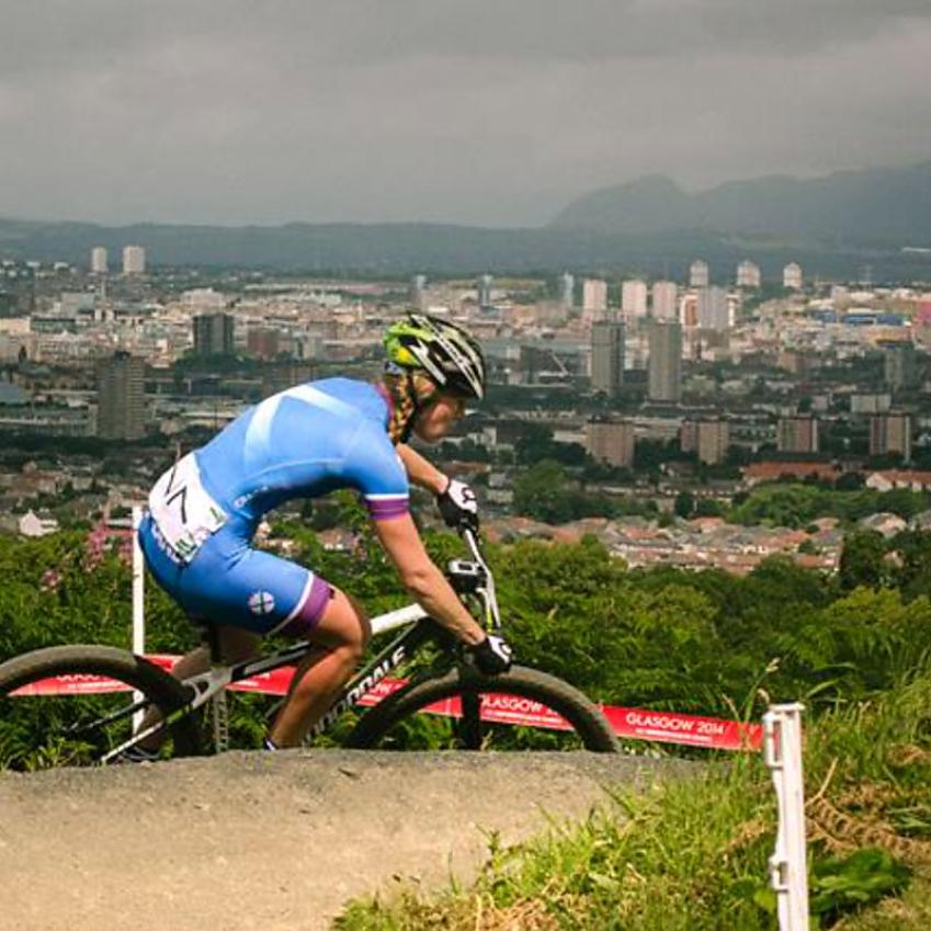 A woman, Lee Craigie, is competing in a mountain bike event in Glasgow. She is wearing blue Scottish cycling gear and riding a black Cannondale mountain bike. The Glasgow skyline is in the background