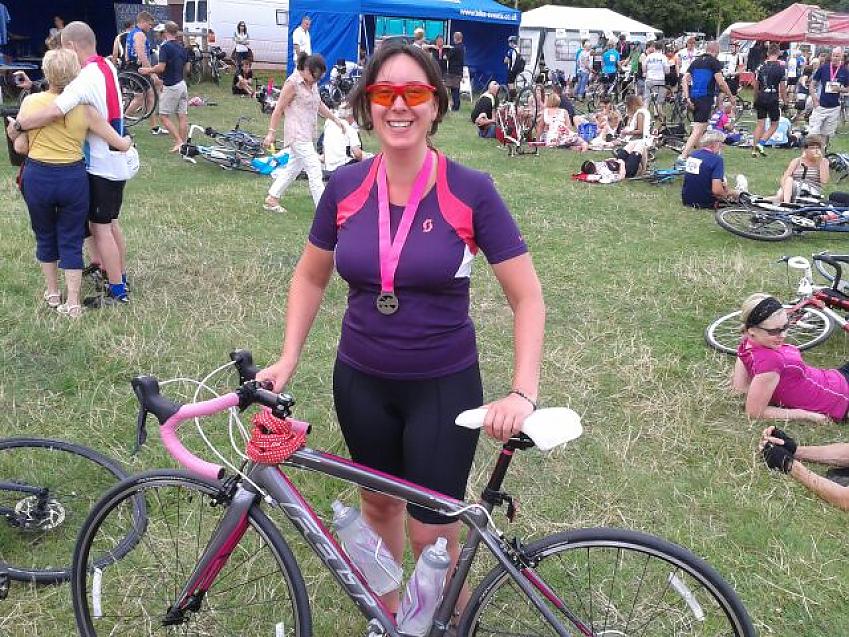 A woman is standing with her road bike in a field. She is wearing cycling kit and has a medal round her neck. She's smiling. There are lots of other cyclists in the background, some lying on the ground with their bikes, and marquees