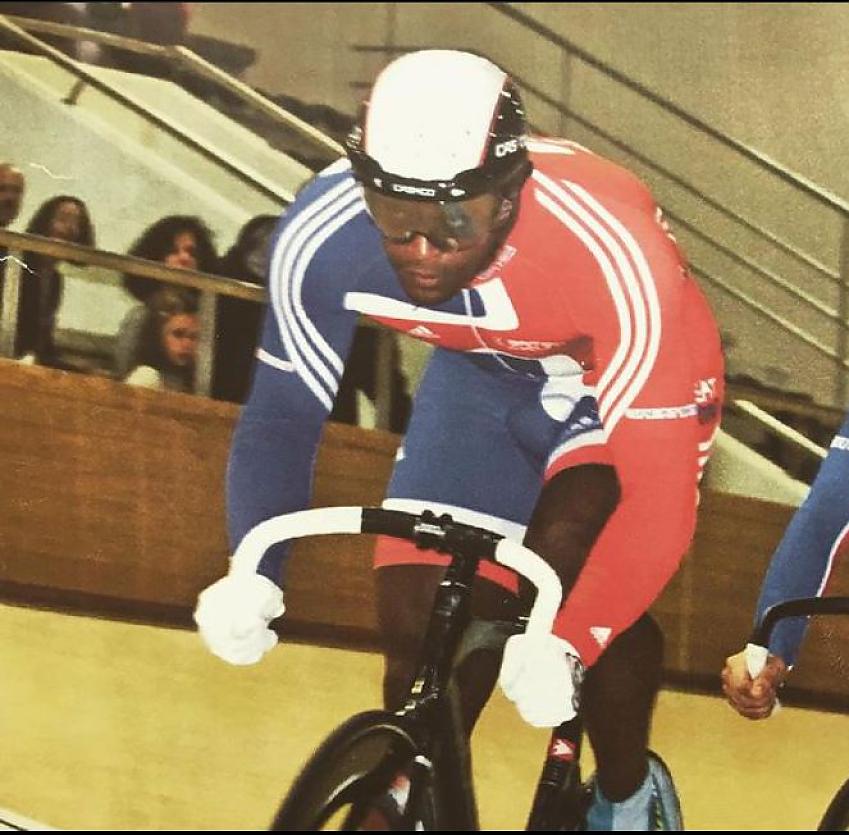 An old photo of a man taking part in a bicycle track race. He is wearing a red, white and blue skin suit.