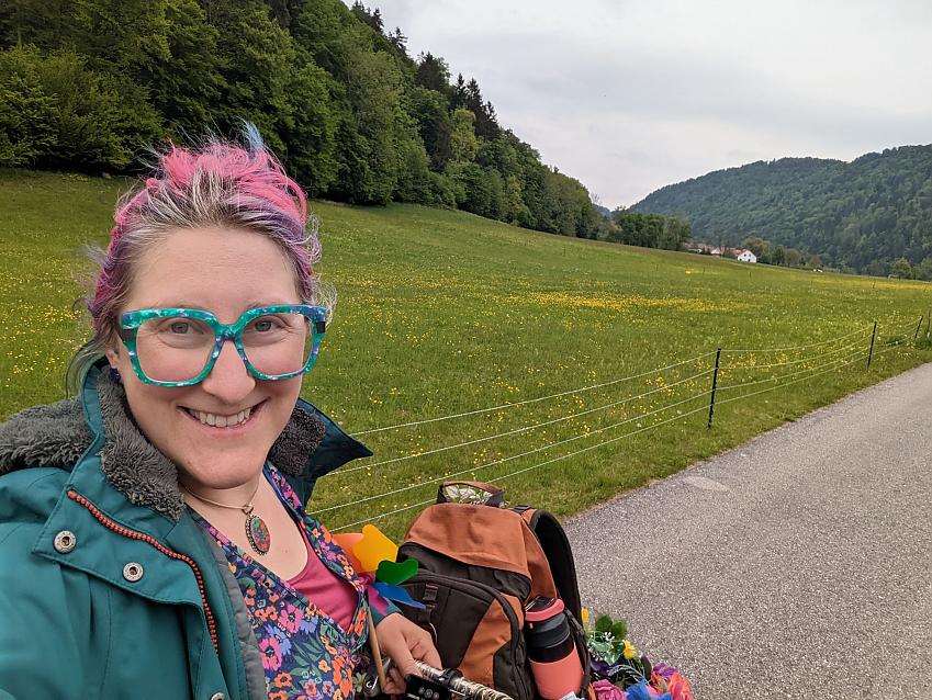 A woman has taken a selfie showing herself and her bike at the top of a hill. She is wearing multicoloured glasses and has multicoloured hair. She is wearing a flowery dress. The luggage in her bike front basket can just be seen.