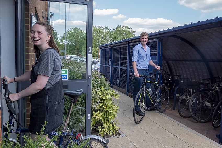 A man is wheeling his bike out of a cycle shed which has lots of cycles in it and places to lock them to. A woman is wheeling her folding bike through the door of a building.