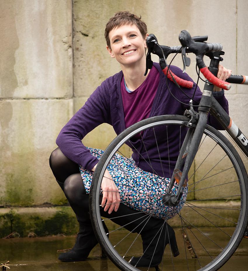 A woman is kneeling behind the front of a road bike. She can be seen through the spokes of the front wheel. She's wearing high-heeled boots, a floral skirt, pink top and purple cardigan. She's smiling