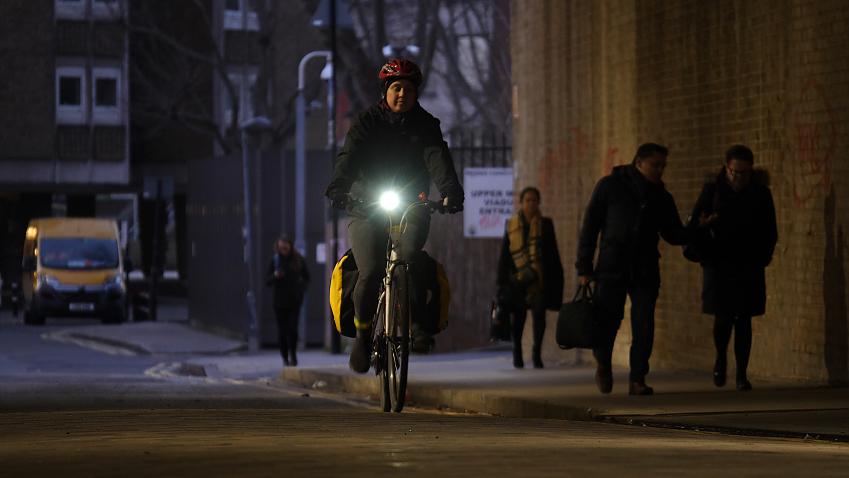 A woman is cycling at dusk in winter. Her bike has two packed panniers. She is wearing winter clothes. She has a bright light on the bike