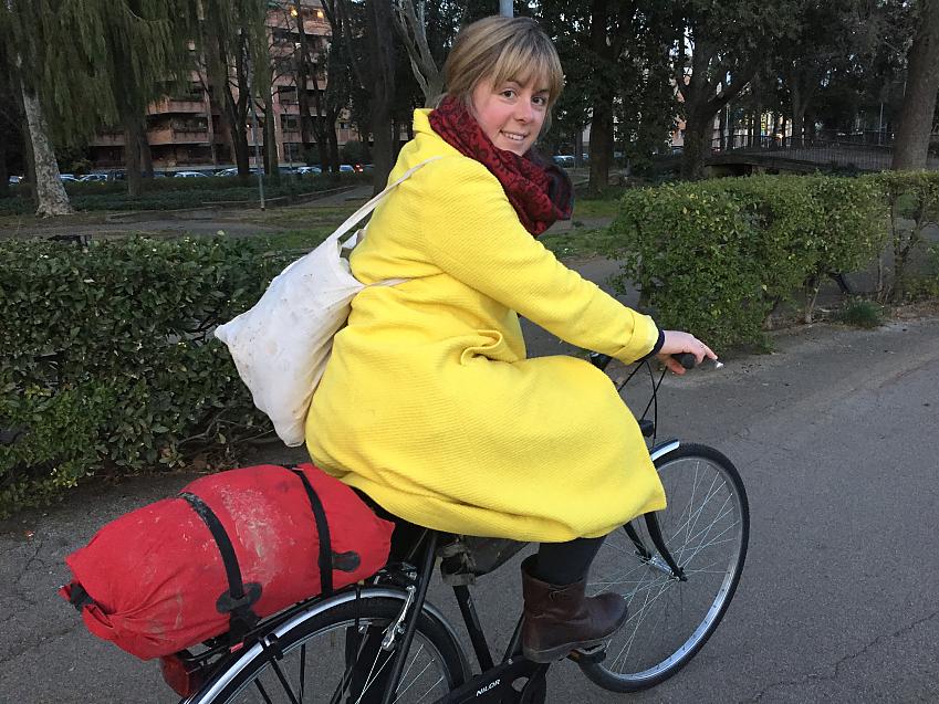 A woman is sitting on her touring bike. She is wearing a bright yellow coat and red and black scarf. She has a white bag over one shoulder and a packed bag on her rear rack